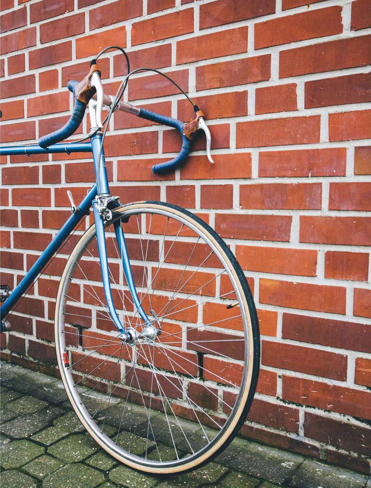 Ausschnitt vom Vorderreifen und Lenker eines blau lackierten Rennrads vor einer roten Backsteinfassade.